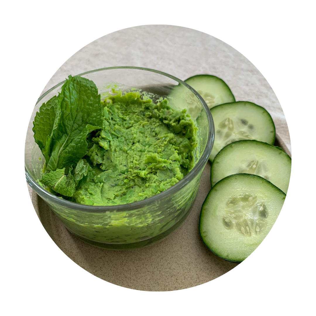 Clear glass container holding green pea dip garnished with mint leaves, on top of a grey plat of four cucumber slices